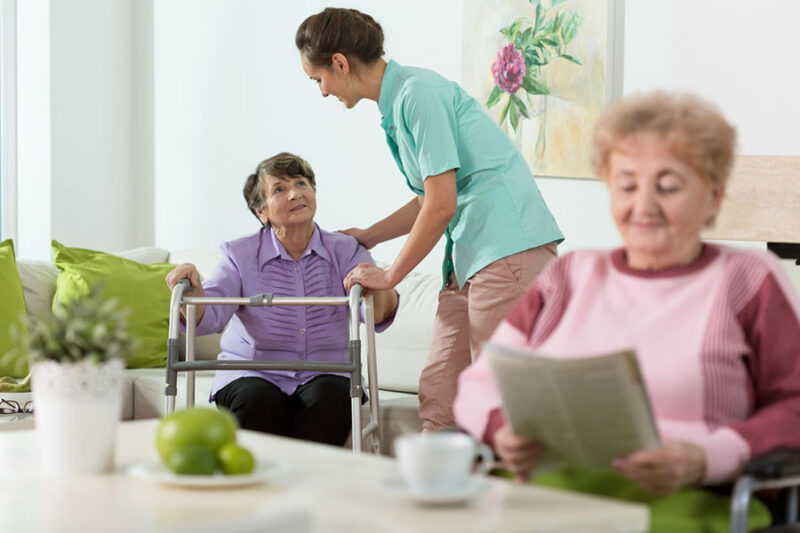 An elderly woman uses a walker with caregiver support, while another reads, showing what is the difference between assisted living and a nursing home.