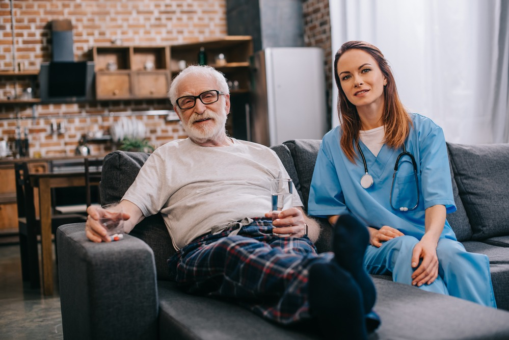 Elderly man relaxing on a sofa with a nurse in scrubs beside him, illustrating senior assisted living in a home-like setting