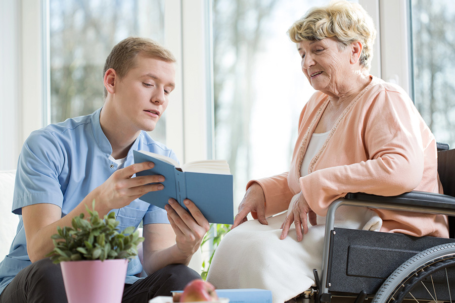 A male caregiver reading to a senior woman in a wheelchair, supporting those with age associated memory impairment