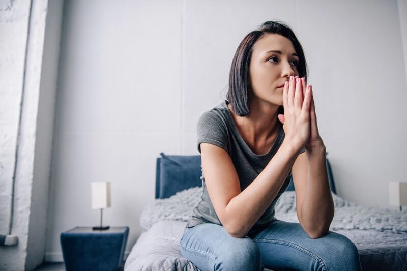 A woman sitting on a bed with a worried expression, illustrating the emotional toll of caregiver burnout