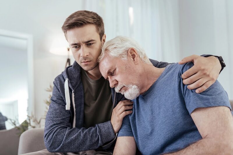 A younger man comforting an older man who appears emotionally exhausted, depicting caregiver fatigue