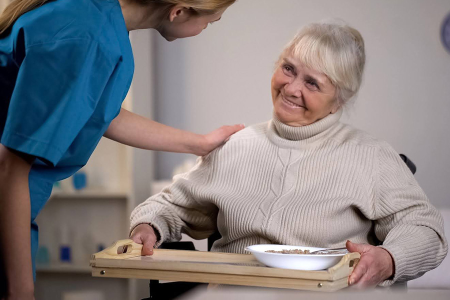 Caregiver offering a meal to a smiling senior woman in a wheelchair, highlighting top assisted living features in Maryland. Caregiver offering a meal to a smiling senior woman in a wheelchair, highlighting top assisted living features in Maryland.
