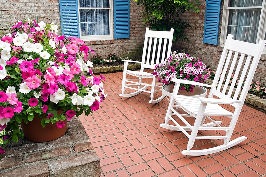 White rocking chairs on a brick patio with vibrant flowers, showcasing a calm outdoor space for senior memory care