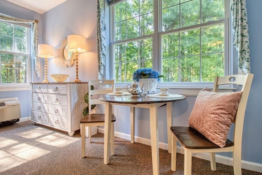 A bright, private studio apartment with a small dining table next to a window, showing the comfortable layout of an assisted living facility.