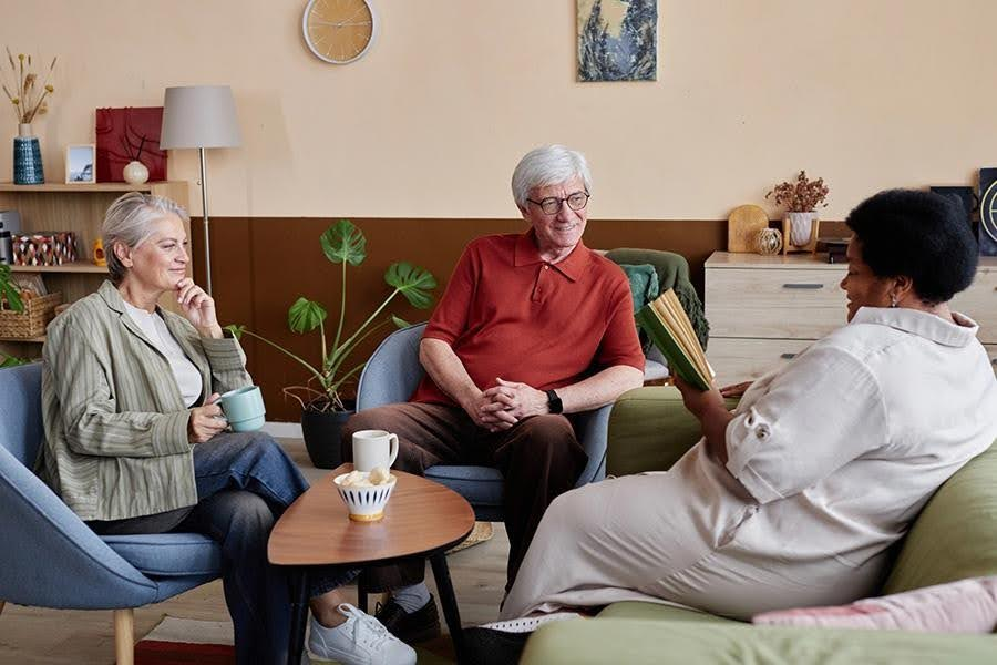 A caregiver reads a book to two seniors in a cozy living room, illustrating the intimate social environment of a small assisted living community.