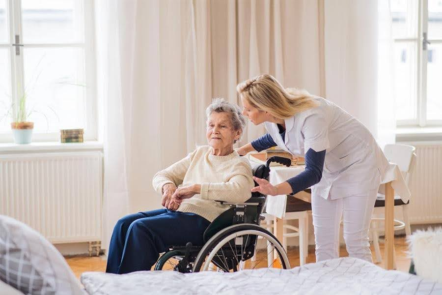 A nurse assists an elderly woman in a wheelchair in her private room, demonstrating the personalized mobility support available in a smaller senior living community.