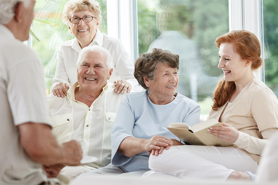 A smiling caregiver reads a book to a group of happy seniors in a bright, sunlit room at an assisted living near Selbyville, DE.