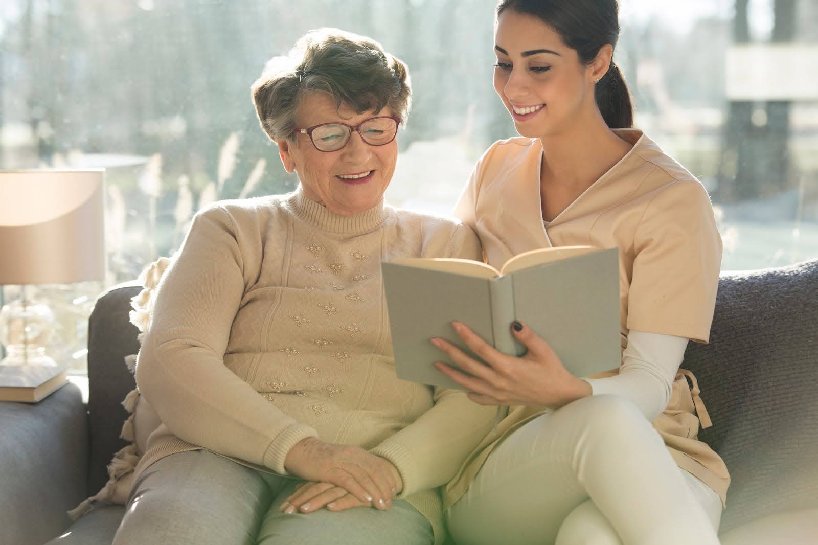 Caregiver reading with elderly woman on a sofa, showing comfort and support during the process of moving to memory care. Caregiver reading with elderly woman on a sofa, showing comfort and support during the process of moving to memory care.
