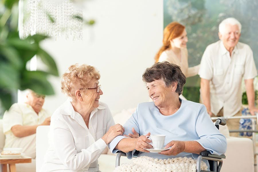 Two senior women smiling and chatting at an assisted living near South Bethany, DE community as a caregiver helps a resident.