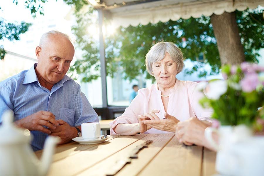 Two seniors enjoying a game of dominoes and coffee on a shaded outdoor patio at an assisted living community